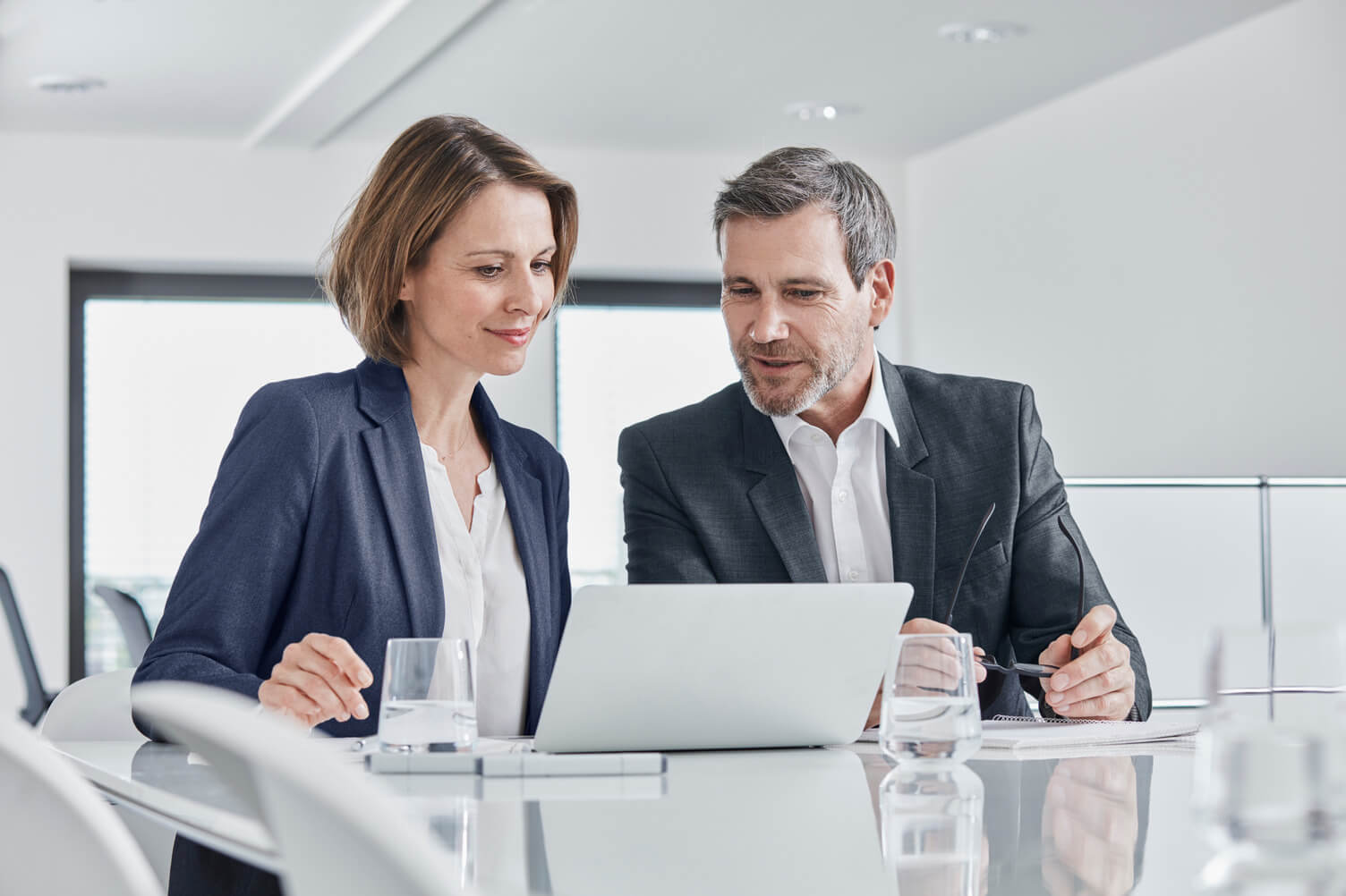 Two business professionals sit at a glass conference table reviewing a laptop in a bright modern office setting.