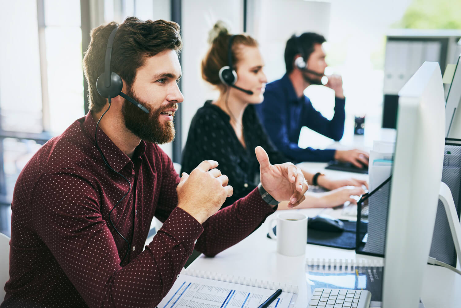 Group of adults wearing headsets work at desktop computers in a modern office, with one man in the foreground speaking while viewing his monitor, representing employer services support, workforce operations, and employment verification processing through Experian Verify™.