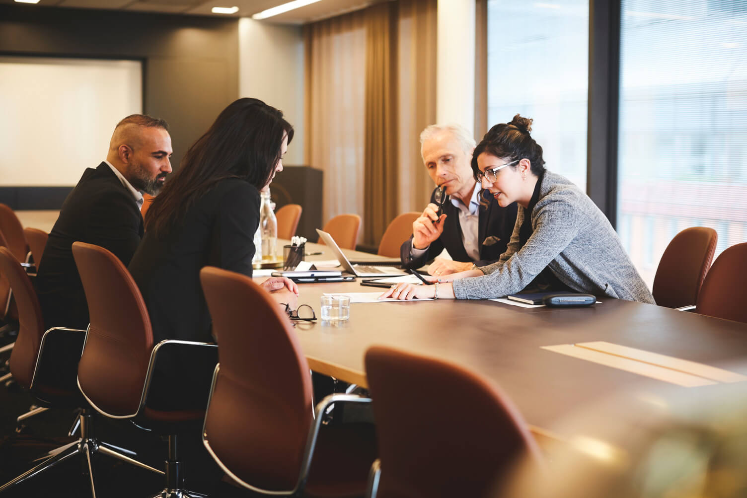 A group of professionals seated around a conference table in a modern office meeting room, engaged in discussion and collaboration. Several people are using laptops and taking notes, with large windows and warm lighting in the background. The image is softly focused, creating a professional and business-oriented atmosphere.