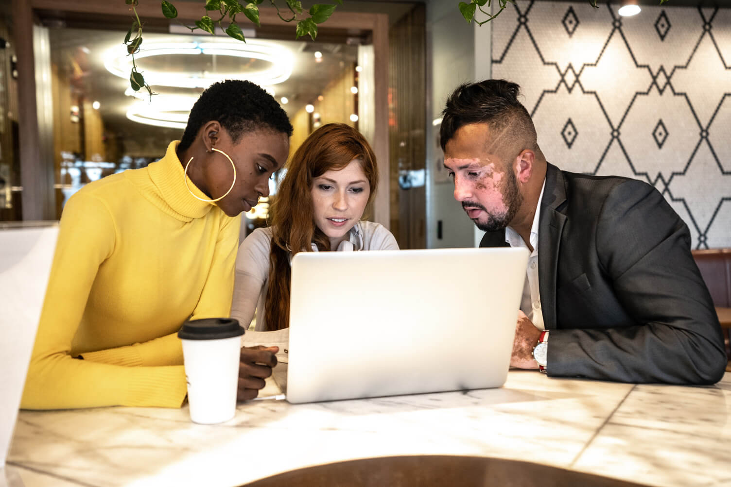 Three people sitting at a marble table in a modern café, focused on a laptop screen, attending an unemployment hearing, with a coffee cup nearby and patterned wall in the background.