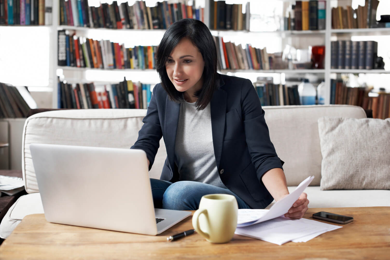 Asian woman in business attire sits at a table with a laptop and papers, working in a home office with bookshelves in the background.