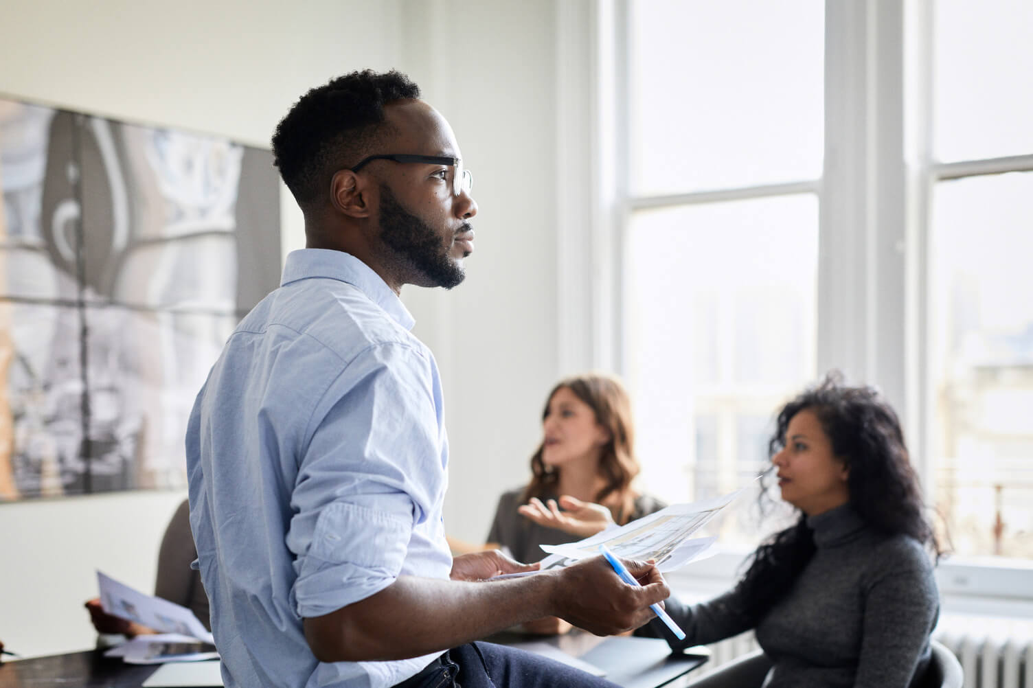 Black man in glasses stands in a bright office holding documents, speaking to two women seated at a conference table during a business meeting.