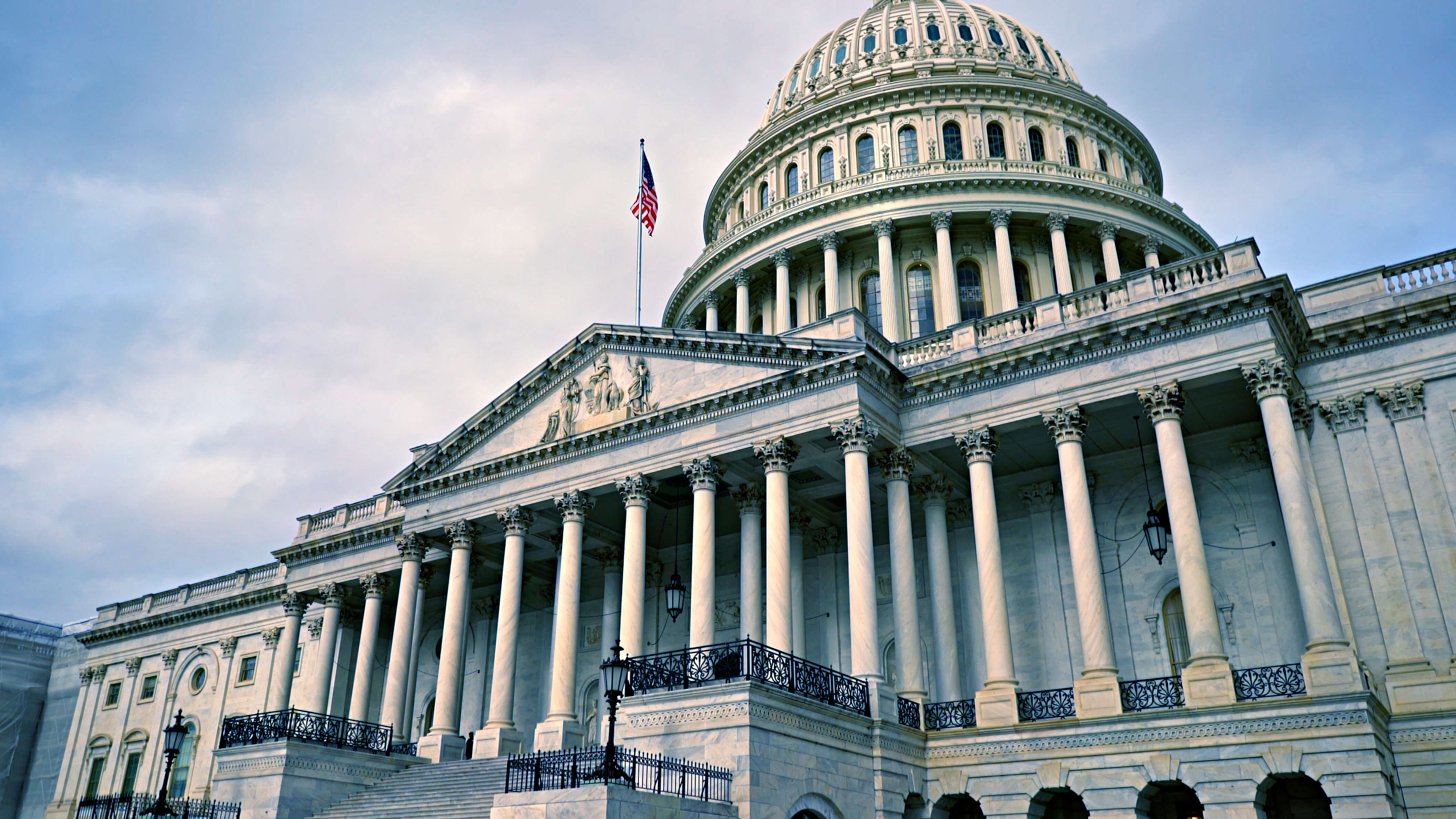 Exterior view of the U.S. Capitol building with its white dome and columns, seen from below against a partly cloudy blue sky.