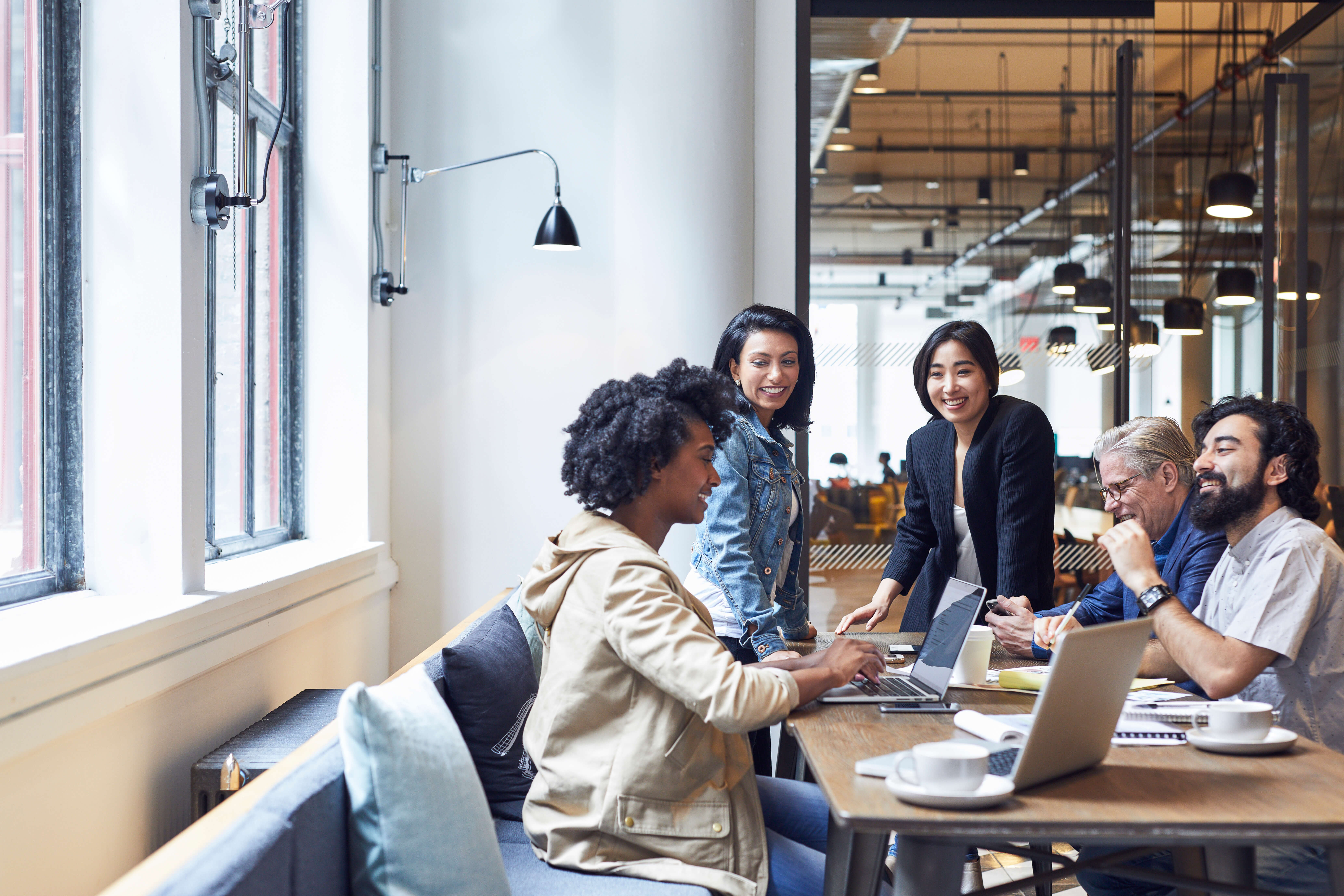 Diverse group of adults collaborate at a long table in a modern office, using laptops and documents in a bright, open workspace.